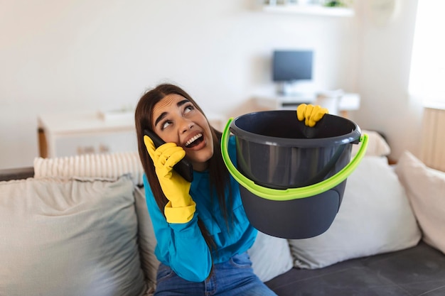 Mujer sorprendida llamando al plomero mientras recolecta agua que se escapa del techo usando un utensilio