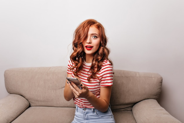 Mujer sorprendida de ojos azules con labios rojos sentada en el sofá. Encantadora chica pelirroja con teléfono posando en su habitación en el sofá.