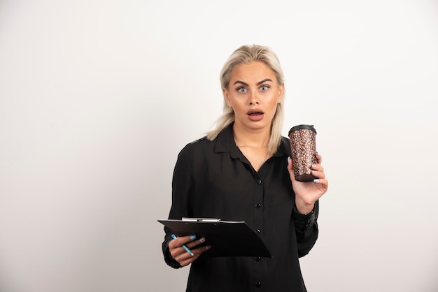 Mujer sorprendida posando con una taza y portapapeles sobre fondo blanco. Foto de alta calidad