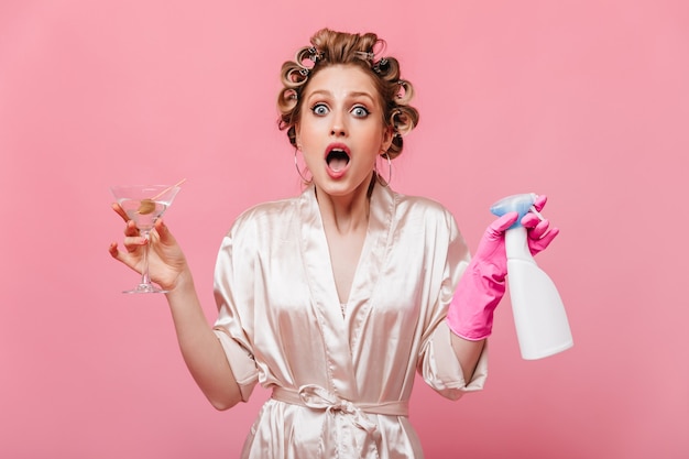 Mujer sorprendida con rizadores de pelo mira al frente y sostiene una copa de martini, herramienta de limpieza