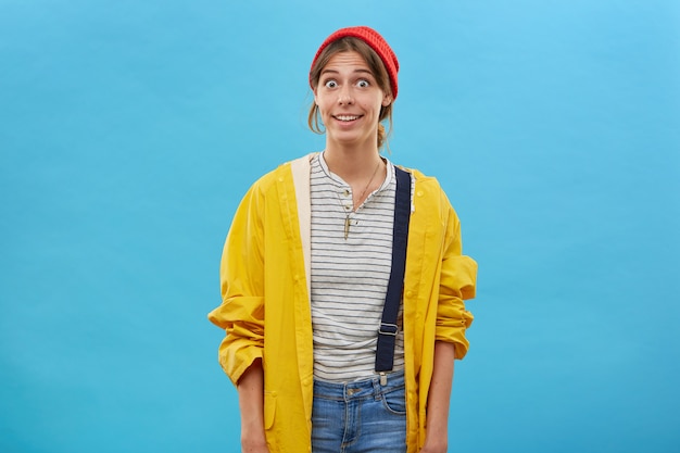 Mujer sorprendida con ropa casual posando contra la pared azul mirando con ojos saltones preguntándose. Mujer joven en impermeable amarillo suelto y sombrero rojo con mirada emocionada