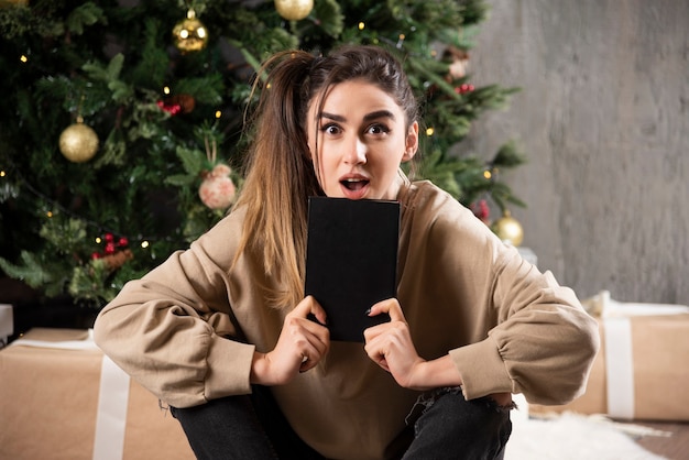 Mujer sorprendida sentada con cuaderno negro cerca del árbol de Navidad.