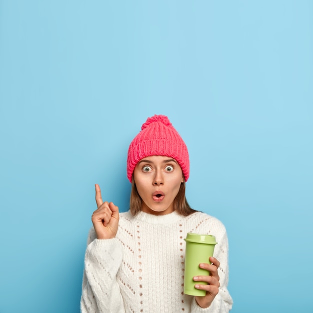 Mujer sorprendida y sorprendida señala con el dedo índice, lleva café para llevar, asustada por algo, mantiene los ojos abiertos