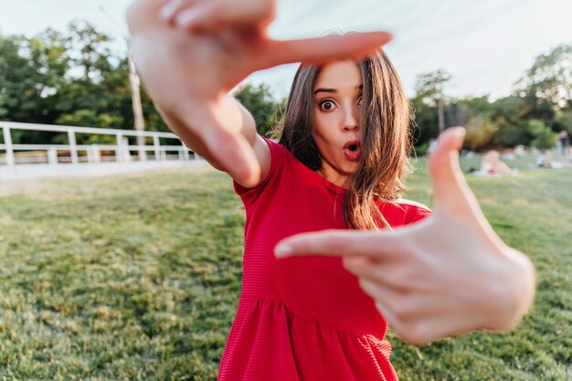 Mujer sorprendida en traje rojo posando al aire libre y haciendo muecas. Foto de niña de cabello castaño emocional escalofriante en primavera.