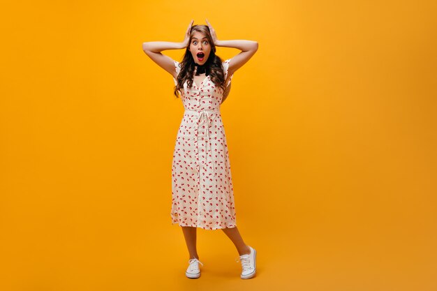 Mujer sorprendida en vestido midi mira a la cámara. Chica rizada en traje de verano blanco y zapatillas posando sobre fondo naranja.
