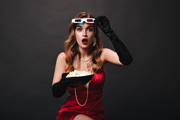 Mujer sorprendida en vestido rojo quitándose gafas 3D y sosteniendo palomitas de maíz Mujer sorprendida en traje festivo de seda viendo una película sobre fondo negro