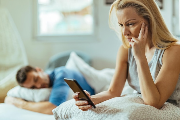 Mujer sospechosa usando el teléfono móvil de su novio mientras él duerme en la cama detrás de ella.