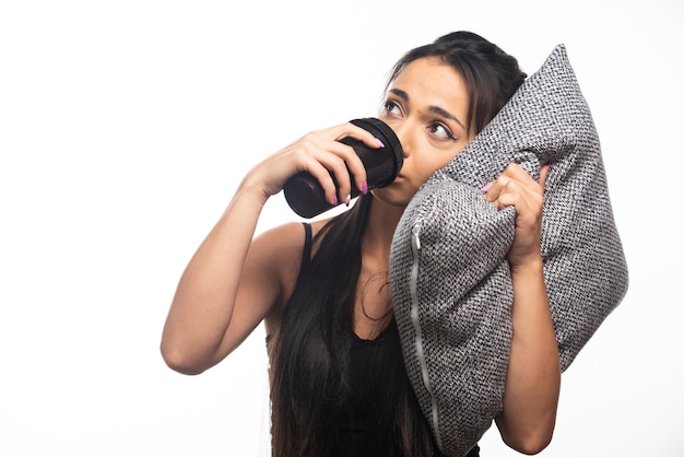 Mujer sosteniendo una almohada y bebiendo de una taza en la pared blanca