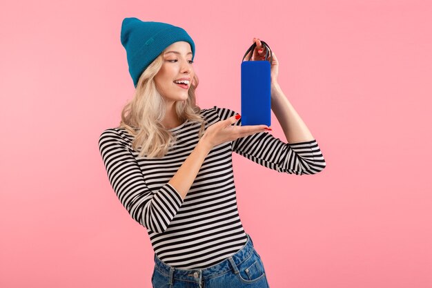 Mujer sosteniendo altavoz inalámbrico escuchando música vistiendo camisa a rayas y sombrero azul sonriendo feliz estado de ánimo positivo posando en rosa