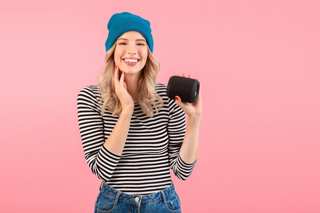 Mujer sosteniendo altavoz inalámbrico escuchando música vistiendo camisa a rayas y sombrero azul sonriendo feliz estado de ánimo positivo posando en rosa