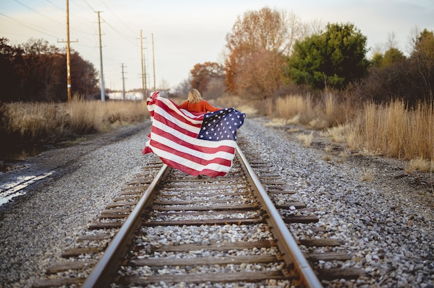 Mujer sosteniendo la bandera americana mientras camina sobre el ferrocarril