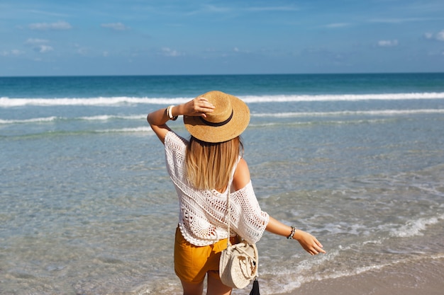 Mujer sosteniendo una bolsa de paja y caminando por la playa