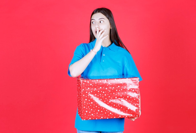 mujer sosteniendo una caja de regalo roja con puntos blancos y parece sorprendida y aterrorizada.