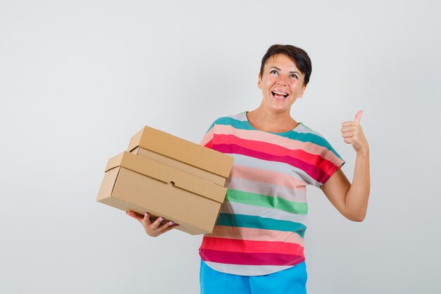 Mujer sosteniendo cajas de cartón, mostrando el pulgar hacia arriba en camiseta a rayas, pantalones y luciendo feliz.