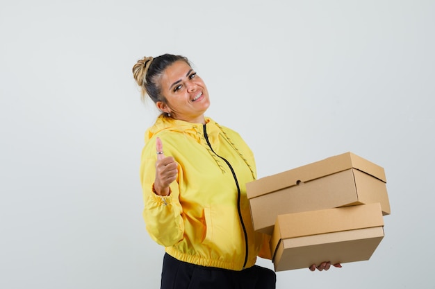 Foto gratuita mujer sosteniendo cajas de cartón, mostrando el pulgar hacia arriba en traje deportivo y luciendo confiada. vista frontal.