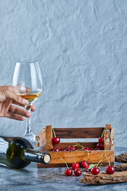 Mujer sosteniendo una copa de vino blanco y caja de madera de cerezas sobre superficie azul