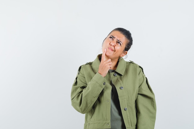 Mujer sosteniendo el dedo en la barbilla en chaqueta, camiseta y mirando vacilante.