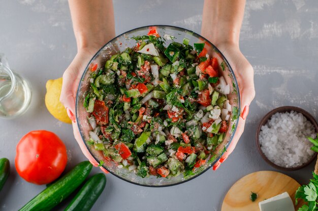 Mujer sosteniendo ensalada de verduras con tomate, queso, pepino, sal en un recipiente de vidrio sobre una superficie gris, vista superior