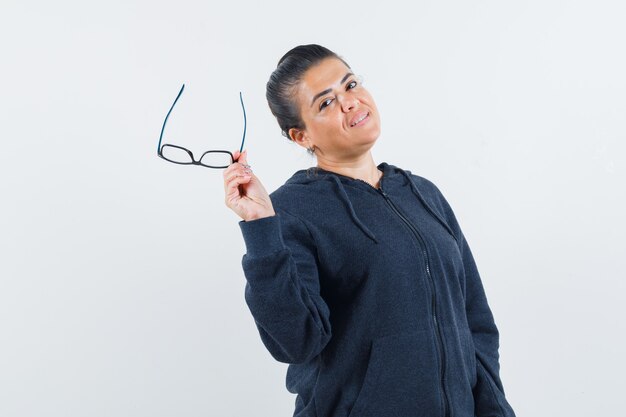 Mujer sosteniendo gafas en sudadera con capucha y mirando confiado. vista frontal.