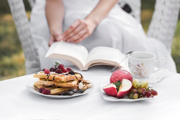 Una mujer sosteniendo un libro en la mano con el desayuno en la mesa blanca en el jardín doméstico