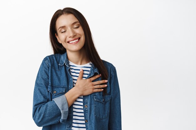 mujer sosteniendo la mano en el corazón, sonriendo nostálgico, pensando en algo romántico o encantador, evocando recuerdos reconfortantes, de pie sobre blanco.