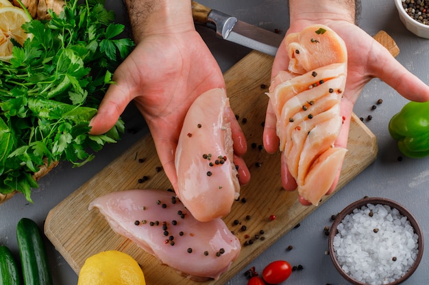 Mujer sosteniendo pechugas de pollo con verduras, pepino, limón, sal vista superior sobre una superficie gris