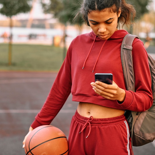 Mujer sosteniendo una pelota de baloncesto al aire libre y revisando su teléfono