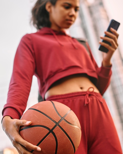 Mujer sosteniendo una pelota de baloncesto al aire libre y revisando su teléfono