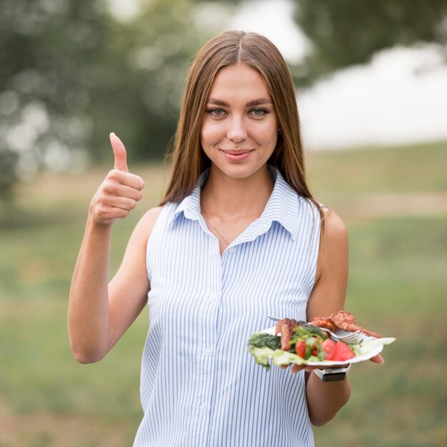 Mujer sosteniendo placa con barbacoa al aire libre y dando pulgar hacia arriba