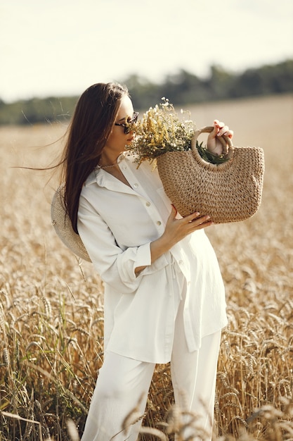 Mujer sosteniendo ramo de flores silvestres en bolsa de paja, caminando en el campo de trigo. Mujer morena caminando en el campo de verano con ropa blanca