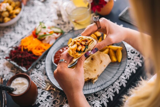 Mujer sosteniendo shawarma de pollo en pan plano servido con papas fritas, salsas, yogur y pepinillos