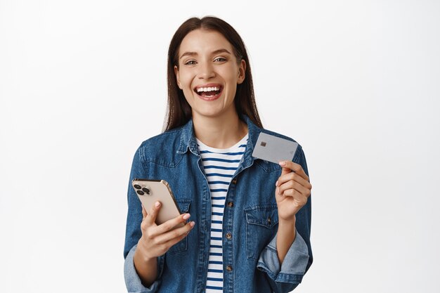 Mujer sosteniendo smartphone, mostrando tarjeta de crédito, sonriendo satisfecho y riendo comprando algo en el teléfono móvil en blanco