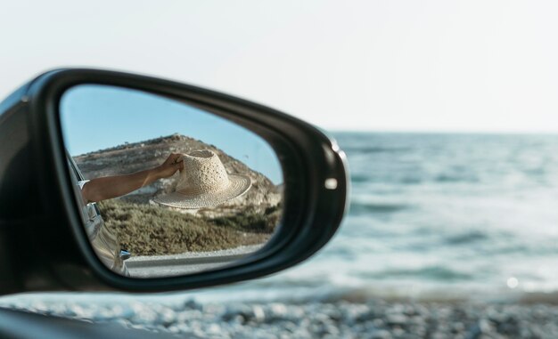 Mujer sosteniendo el sombrero por la ventana en la vista del espejo de coche