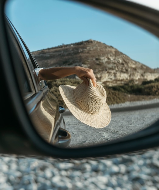 Mujer sosteniendo el sombrero por la ventana en la vista del espejo de coche