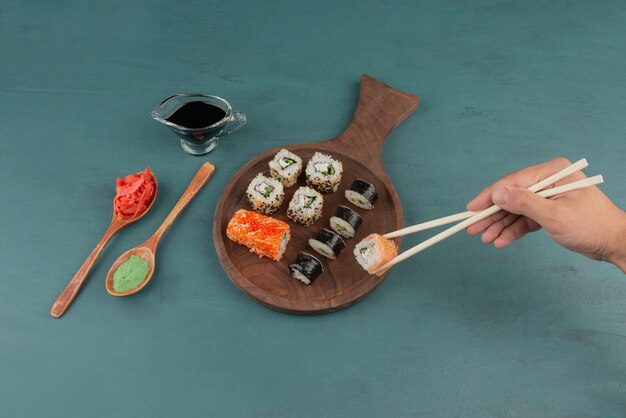 Mujer sosteniendo sushi roll con palillos en la mesa azul con jengibre encurtido y salsa de soja.