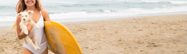 Mujer sosteniendo una tabla de surf y un perro en la playa.