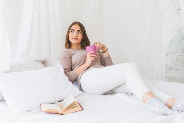 Mujer sosteniendo la taza de café acostado en la cama con el libro