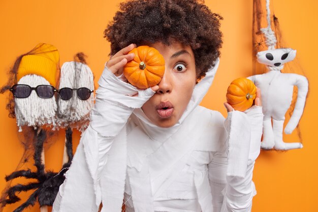 Mujer sostiene dos pequeñas calabazas mira sorprendida viste traje de fantasma blanco rodeado de juguetes de Halloween aislado en naranja