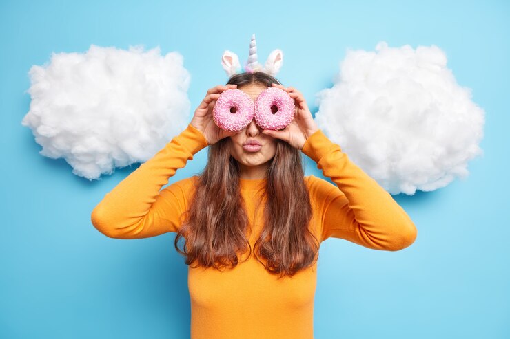 Foto gratuito mujer sostiene dos rosquillas dulces horneadas en los ojos mantiene los labios doblados disfruta comiendo sabrosa comida alta en calorías vestida con un jersey naranja aislado en azul