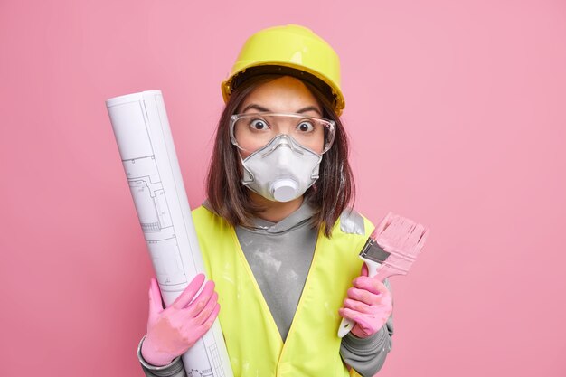 La mujer sostiene el plano y el pincel de pintura usa casco protector, gafas y uniforme trabaja en la construcción y reparación