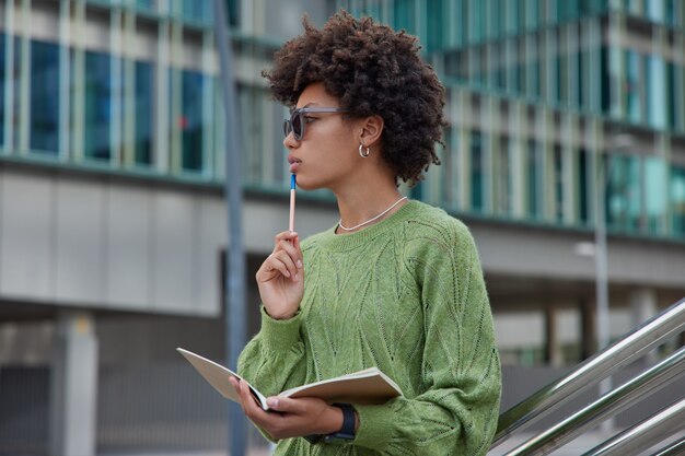 La mujer sostiene la pluma y el diario abierto hace notas programar qué hacer usa gafas de sol puente verde plantea afuera contra el edificio moderno