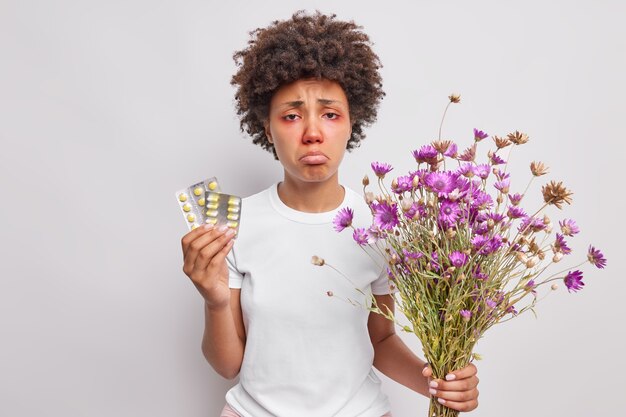 Mujer sostiene un ramo de flores silvestres y pastillas para la alergia tiene ojos rojos y llorosos se ve con expresión triste aislada sobre blanco