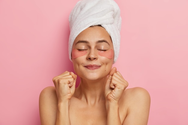 Mujer de spa hace mascarilla para el cabello, aplica parches de hidrogel, aprieta los puños como anticipa el efecto del producto cosmético, se para sin camisa con los ojos cerrados