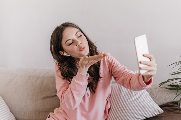 Mujer en sudadera y auriculares inalámbricos sopla beso y toma selfie
