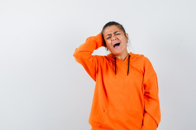 Mujer en sudadera con capucha naranja manteniendo la mano en la cabeza y mirando triste