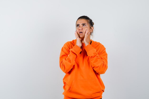 Mujer en sudadera con capucha naranja manteniendo las manos en las mejillas y mirando enfocado