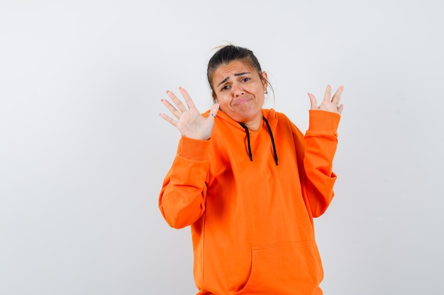 Mujer en sudadera con capucha naranja mostrando las palmas en gesto de rendición y mirando confundido