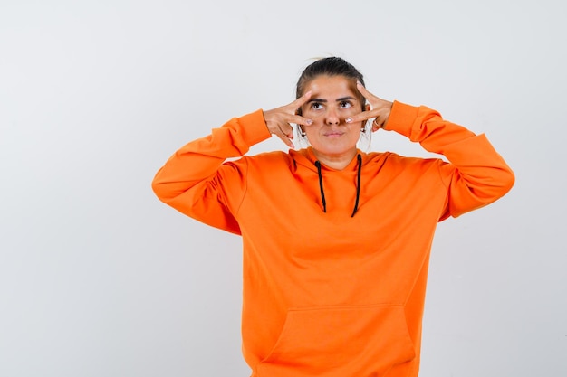 Mujer en sudadera con capucha naranja que muestra el signo V en los ojos y parece vacilante