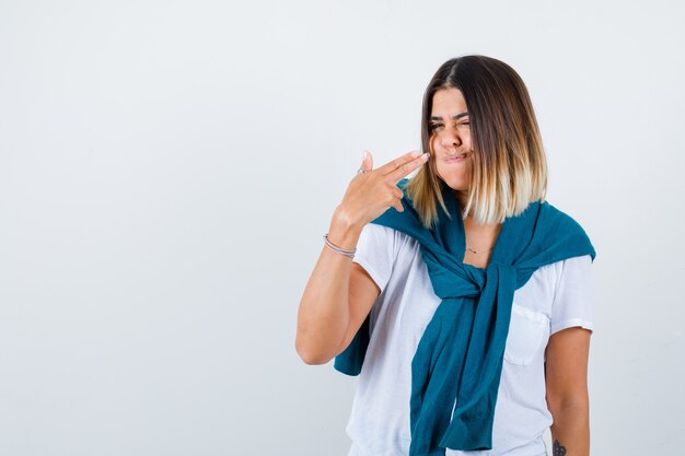 Mujer con suéter atado mostrando gesto de pistola, soplando mejillas en camiseta blanca y luciendo confiada. vista frontal.