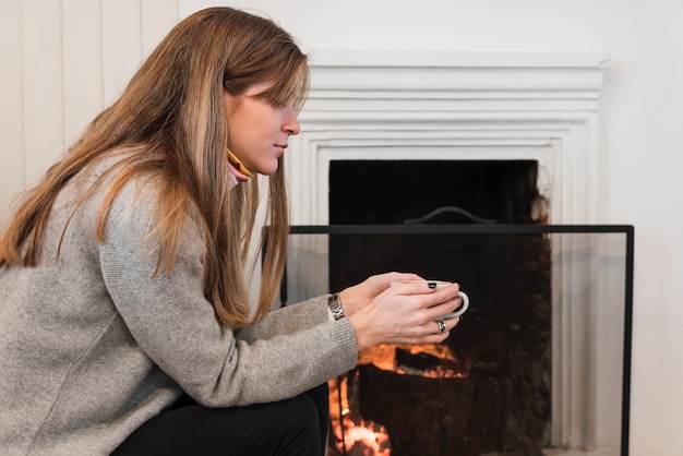 Mujer en suéter bebiendo té junto a la chimenea
